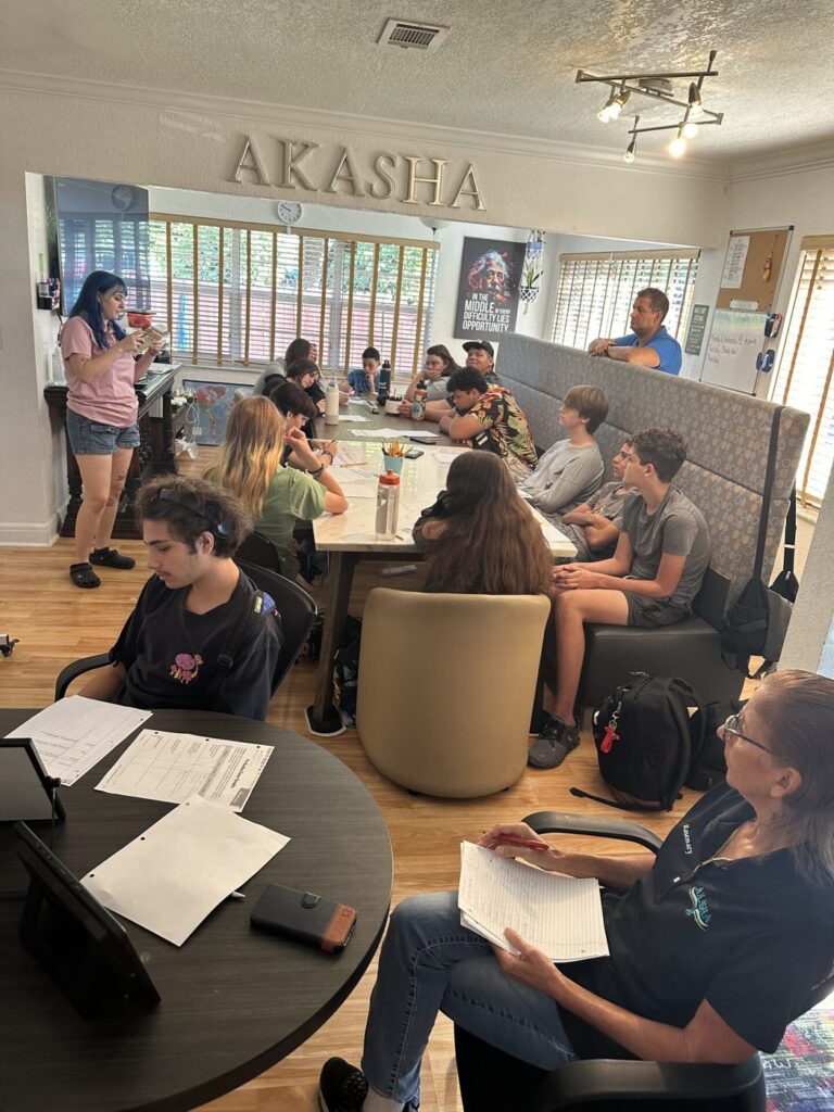 A full room of students working at tables under the AKASHA letters on the wall