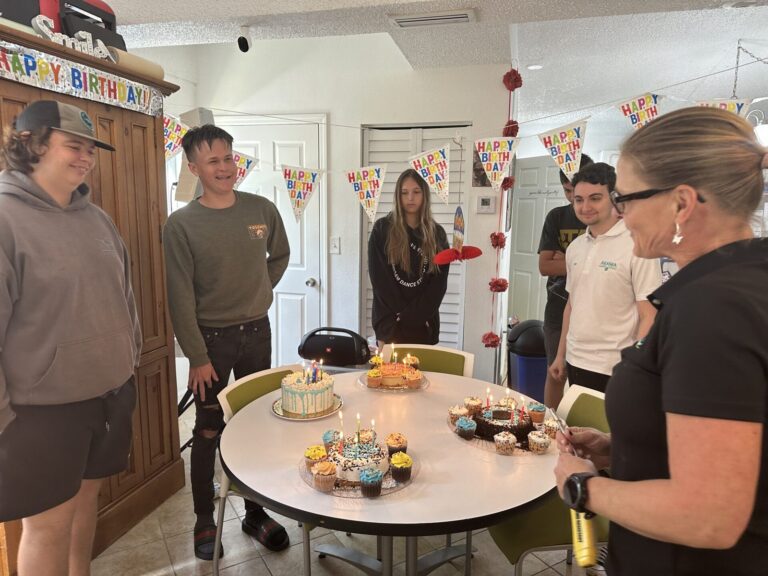 Students and a teacher gather around a table of birthday cake and cupcakes under Happy Birthday banners