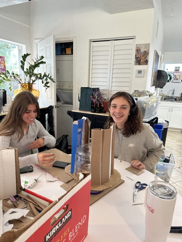 Two smiling students build tall cardboard models at a table