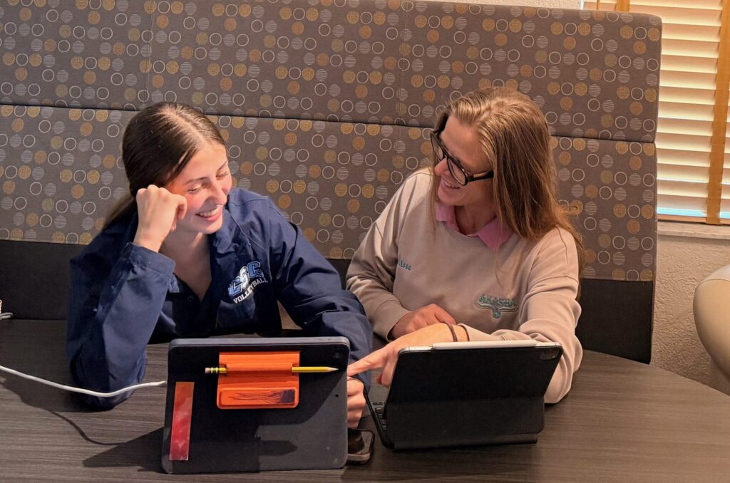 Two students in Akasha shirts laugh together at a booth table while working on iPads