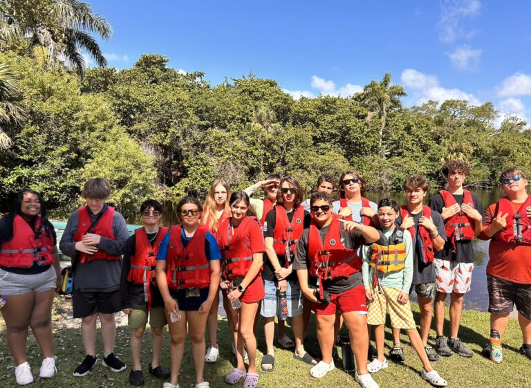 A big group of students in red life jackets lines up for a photo under tropical trees
