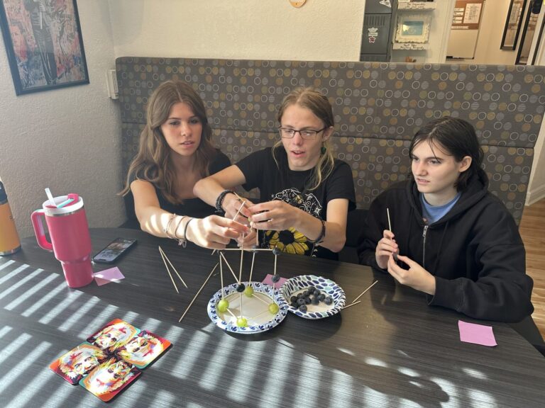 Three students at a table build a wire and straw structure together