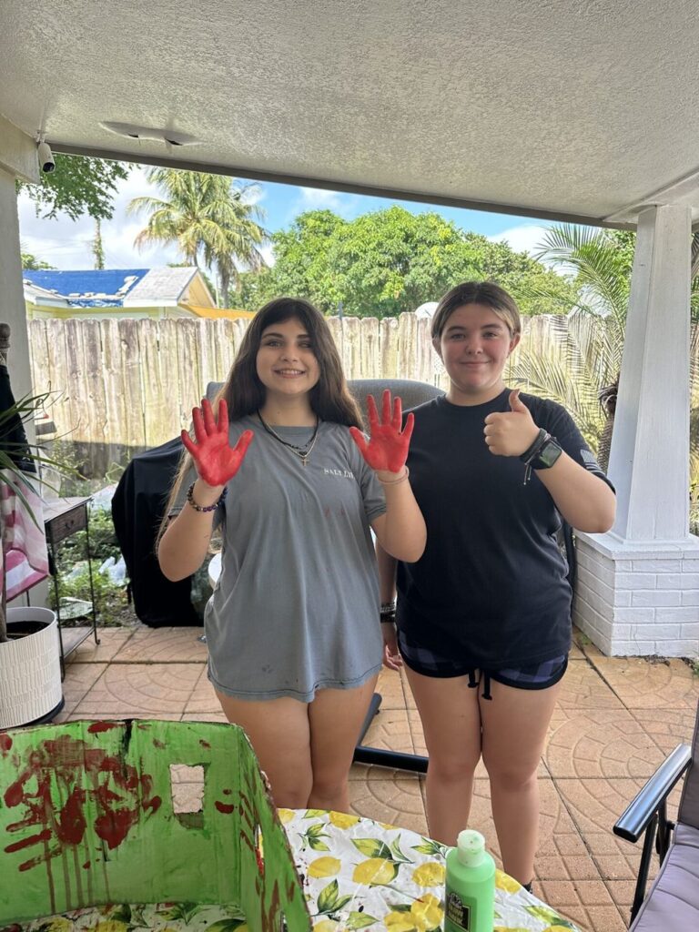 Two students grin and hold up paint-covered red hands next to their art project